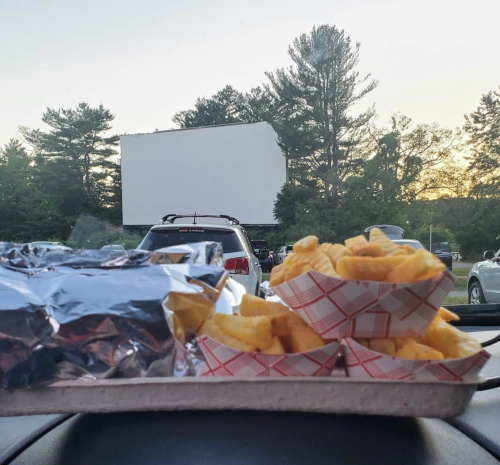 A tray with wrapped food and fries in front of a drive-in movie screen at sunset.