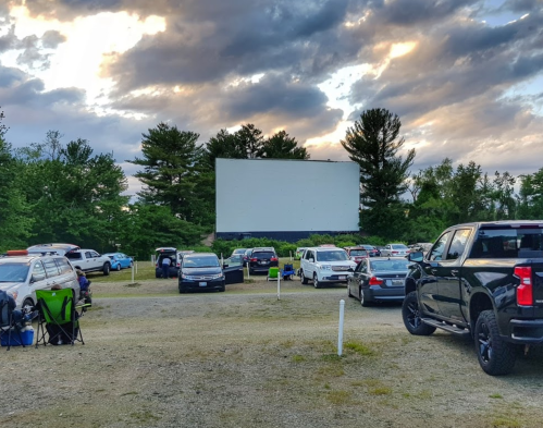 A drive-in movie theater with cars parked in front of a large blank screen, surrounded by trees and cloudy skies.
