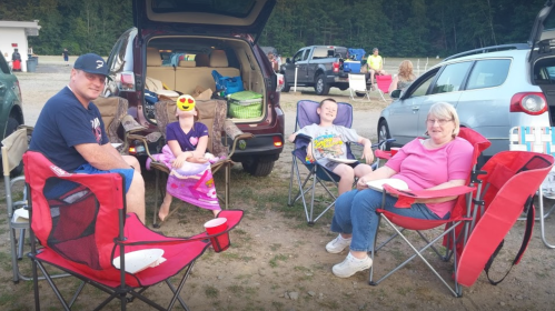 A family sits in camping chairs by their vehicle, enjoying a day outdoors with smiles and relaxed expressions.