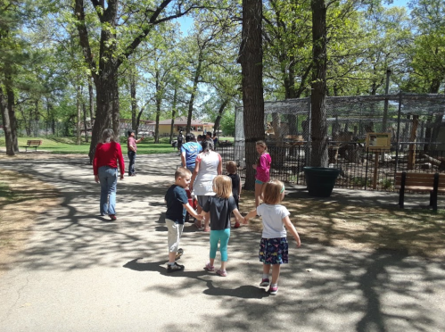 A group of children and adults walking along a path in a park, with trees and a zoo enclosure in the background.