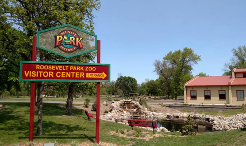 Sign for Roosevelt Park Zoo Visitor Center with a pond and greenery in the background on a sunny day.