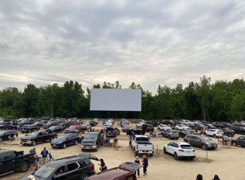 A drive-in movie theater with numerous parked cars and a large blank screen surrounded by trees under a cloudy sky.