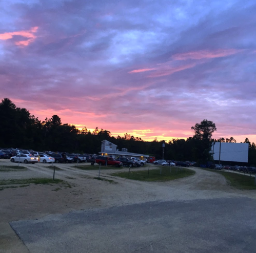 A drive-in movie theater at sunset, with cars parked and a colorful sky filled with pink and purple hues.