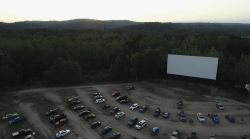 Aerial view of a drive-in movie theater with parked cars and a large blank screen surrounded by trees at sunset.