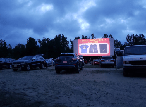A parking lot at dusk with cars and a large screen displaying T-shirts, sweatshirts, and blankets for sale.