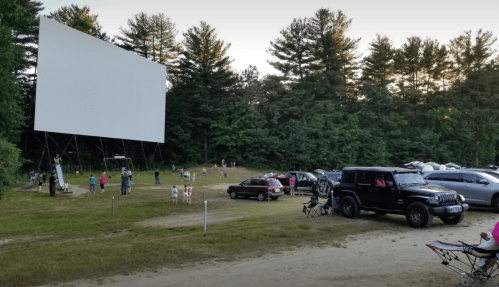 Outdoor movie screening with a large screen, cars parked nearby, and people enjoying the event in a wooded area.