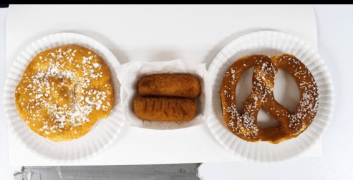 Two paper plates with a funnel cake, two corn dogs, and a soft pretzel, all dusted with powdered sugar.