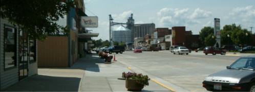 A small town street with shops, parked cars, and a grain silo in the background under a blue sky with clouds.