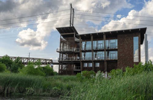 Modern building with large windows, surrounded by greenery and water, under a cloudy sky with power lines in the background.