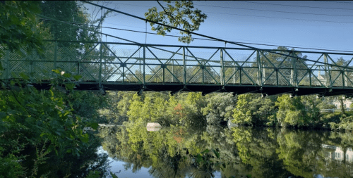 A green pedestrian bridge spans a calm river, surrounded by lush trees and reflecting the clear blue sky.