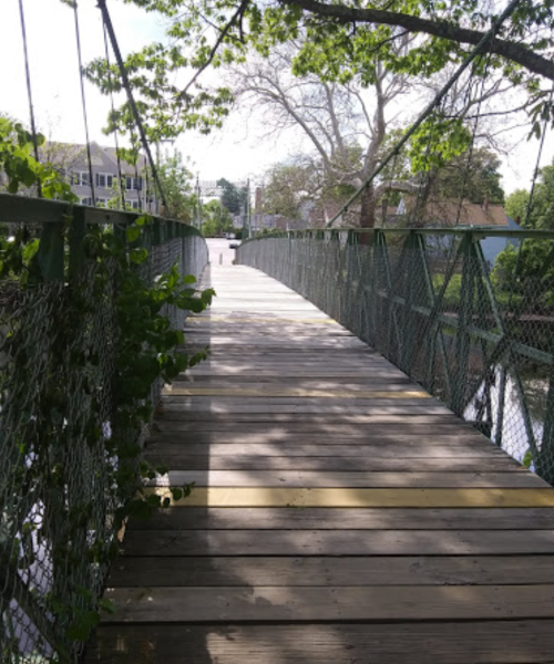 A wooden pedestrian bridge with green railings, surrounded by trees and casting shadows on the walkway.