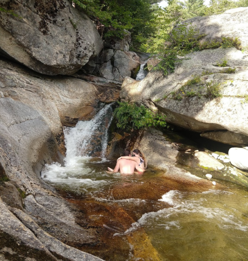 A person sits in a natural pool by a small waterfall, surrounded by large rocks and greenery.