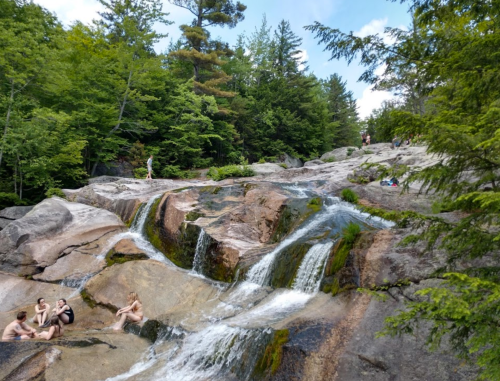 People relax by a rocky waterfall surrounded by lush green trees and a clear blue sky.