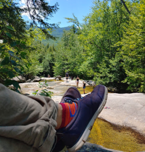 A person's foot in colorful socks rests on a rock by a stream, with people walking in the background among trees.