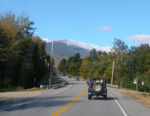 A scenic road winding through trees with a mountain in the background and a black jeep driving ahead.