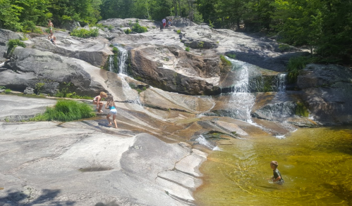 Children play on rocky terrain near a small waterfall and a natural pool surrounded by greenery.