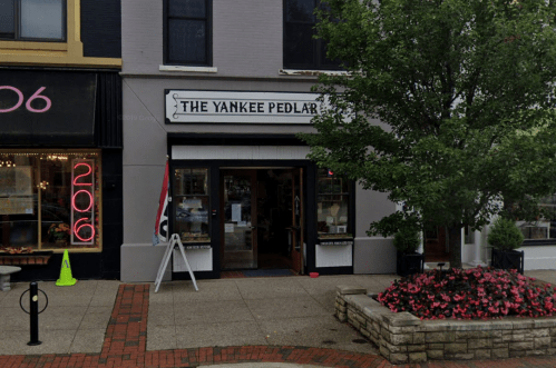 A storefront labeled "The Yankee Pedlar," with a tree and flowers in front, located on a brick sidewalk.