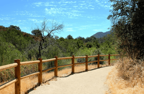 A winding dirt path bordered by a wooden fence, surrounded by lush greenery and distant mountains under a blue sky.