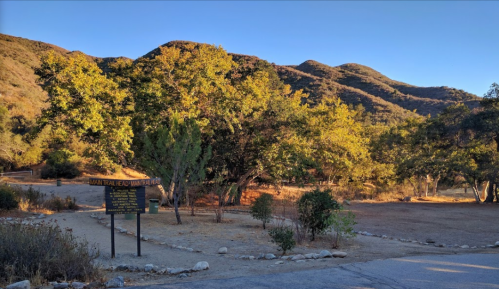 A trailhead sign surrounded by trees and mountains under a clear blue sky in a natural setting.
