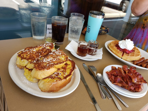 A table with French toast, waffles topped with whipped cream, bacon, and drinks in glasses and a thermos.