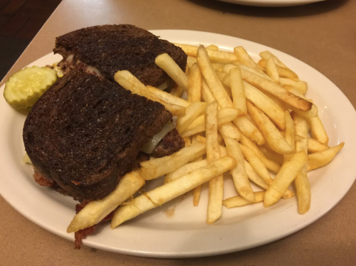 A plate with a grilled sandwich on dark bread, accompanied by a side of golden French fries and a pickle.