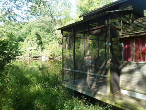 A wooden screened porch overlooks a serene, green landscape by a calm body of water.