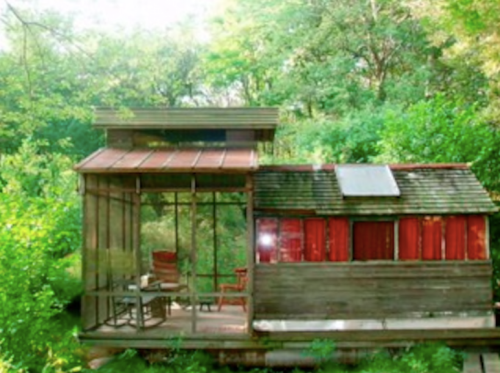 A small wooden cabin surrounded by lush greenery, featuring large windows and a cozy porch.