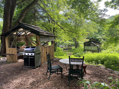 A rustic outdoor space with a grill, table, and chairs surrounded by lush greenery and wooden structures.