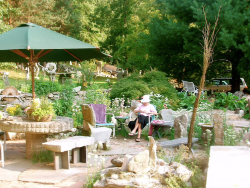 A serene garden scene with stone seating, lush greenery, and a person sitting under an umbrella, enjoying the outdoors.