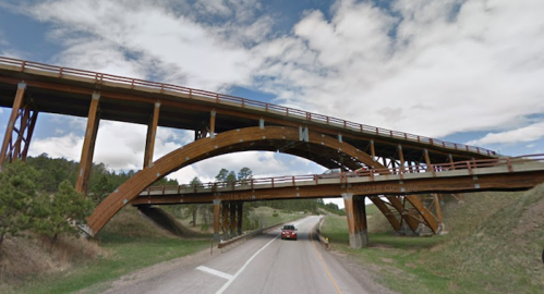 A wooden arch bridge spans over a highway, surrounded by green hills and a cloudy sky.