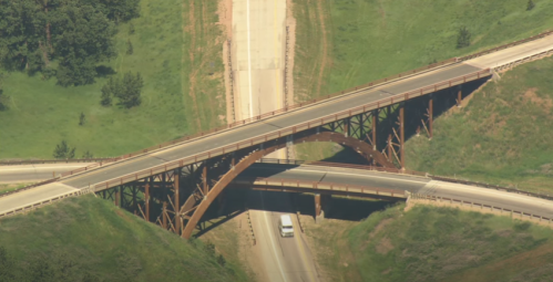 Aerial view of a highway interchange with a curved bridge over intersecting roads surrounded by green hills.