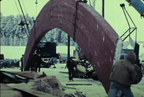 Workers maneuver a large, curved structure at a construction site in winter, surrounded by machinery and materials.