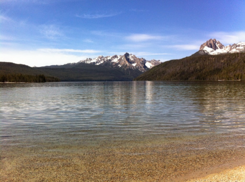 A serene lake surrounded by mountains, with clear water reflecting the blue sky and snow-capped peaks in the distance.