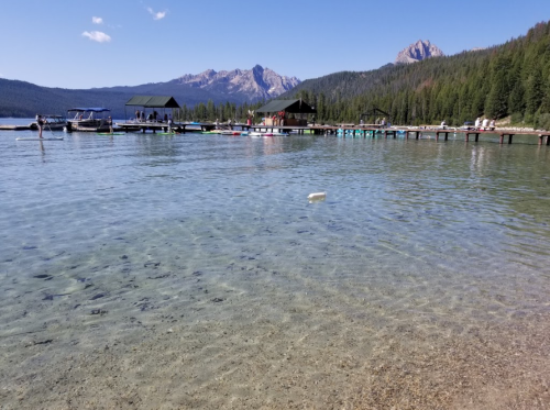 A clear lake with a sandy shore, boats docked, and mountains in the background under a blue sky.