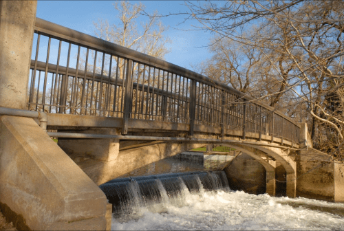 A metal bridge spans a small waterfall, surrounded by trees and a clear blue sky.