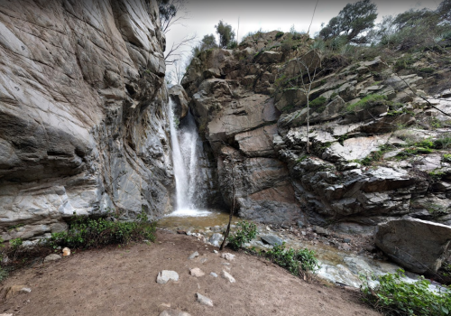 A waterfall cascading down rocky cliffs, surrounded by greenery and a sandy area at the base.