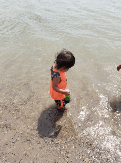 A toddler in an orange outfit stands in shallow water, playing with a green bottle on a sunny day at the beach.