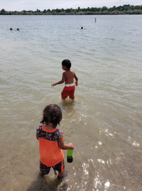 Two children play in shallow water at a lake, one holding a bottle and the other wading in the water.