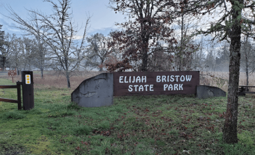 Sign for Elijah Bristow State Park surrounded by trees and grass in a misty landscape.