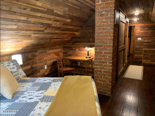 Cozy attic bedroom with wooden walls, a bed, desk, lamp, and a warm, inviting atmosphere.