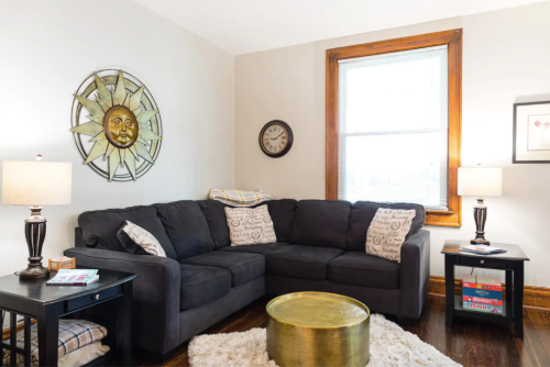 Cozy living room with a dark sectional sofa, decorative sun wall art, lamps, and a gold coffee table.