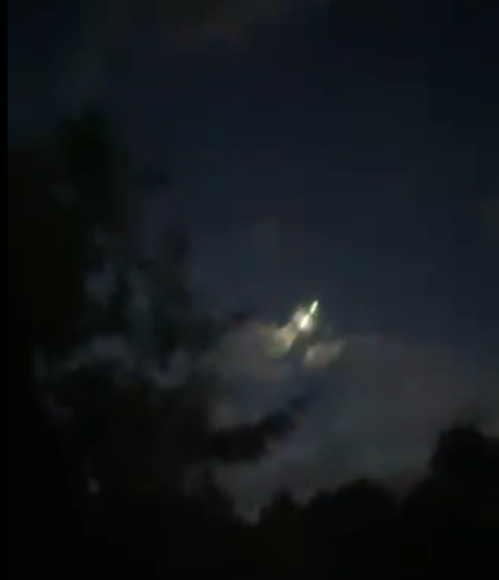 A dimly lit night sky with clouds and a bright light, possibly the moon, partially obscured by trees.