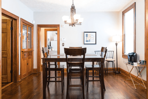 A cozy dining area with a wooden table, chairs, and warm lighting, featuring a lamp and a coffee maker in the corner.