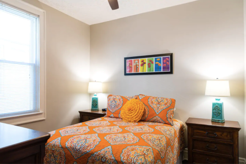 Cozy bedroom with an orange patterned bedspread, two lamps, and colorful artwork on the wall.