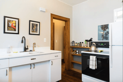A modern kitchen with white cabinets, a black stove, and framed art on the walls, featuring a wooden door and shelves.