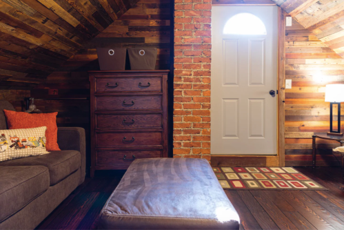 Cozy attic room with a brown sofa, wooden dresser, and a door leading outside, featuring rustic brick and wood walls.