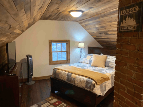 Cozy attic bedroom with wooden beams, a bed, TV, lamp, and a sign pointing to the lodge.