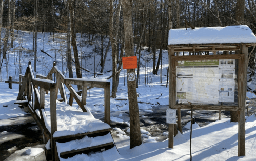 A snowy landscape featuring a wooden bridge over a stream and a trail information sign in a forested area.