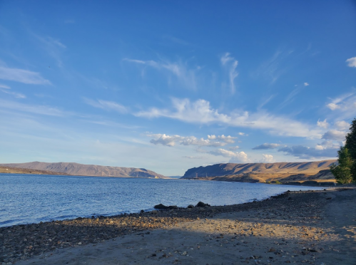 A serene lakeside view with rocky shorelines, mountains in the distance, and a clear blue sky with wispy clouds.