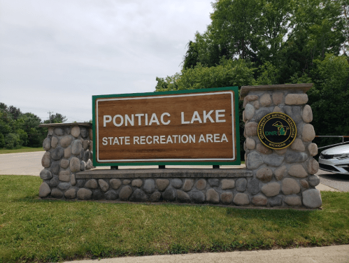 Sign for Pontiac Lake State Recreation Area, featuring a wooden board and stone base, surrounded by greenery.